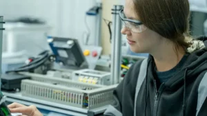 Kobieta w okularach ochronnych przy stanowisku z urządzeniami elektronicznymi i przewodami w hali produkcyjnej